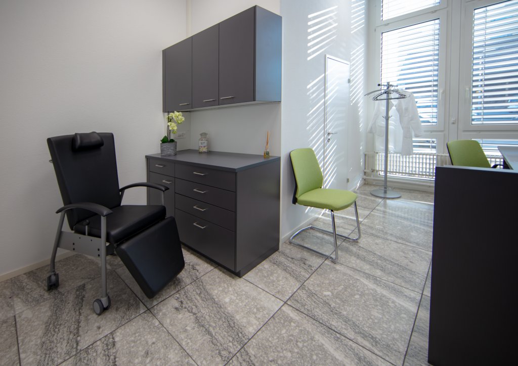 Eine saubere, moderne Arztpraxis am Standort D&auml;ttwil mit schwarzem Lehnstuhl, gr&uuml;nem Stuhl, grauen Schr&auml;nken und einer Garderobe mit einem weissen Laborkittel neben grossen Fenstern mit Jalousien.