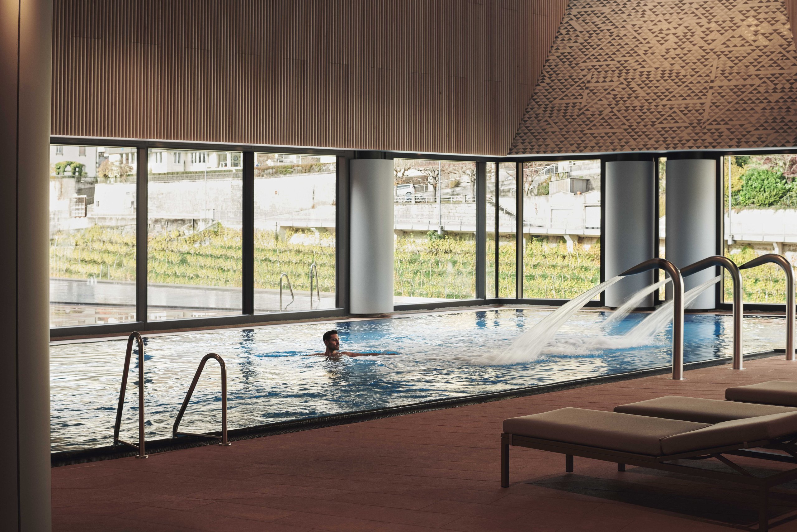 Eine Person genie&szlig;t Wellness in einem modernen FORTYSEVEN-Hallenbad mit Wasserd&uuml;sen, gro&szlig;en Fenstern mit Blick ins Freie und Liegest&uuml;hlen in der N&auml;he. Nat&uuml;rliches Licht erf&uuml;llt den ruhigen, ger&auml;umigen Bereich.