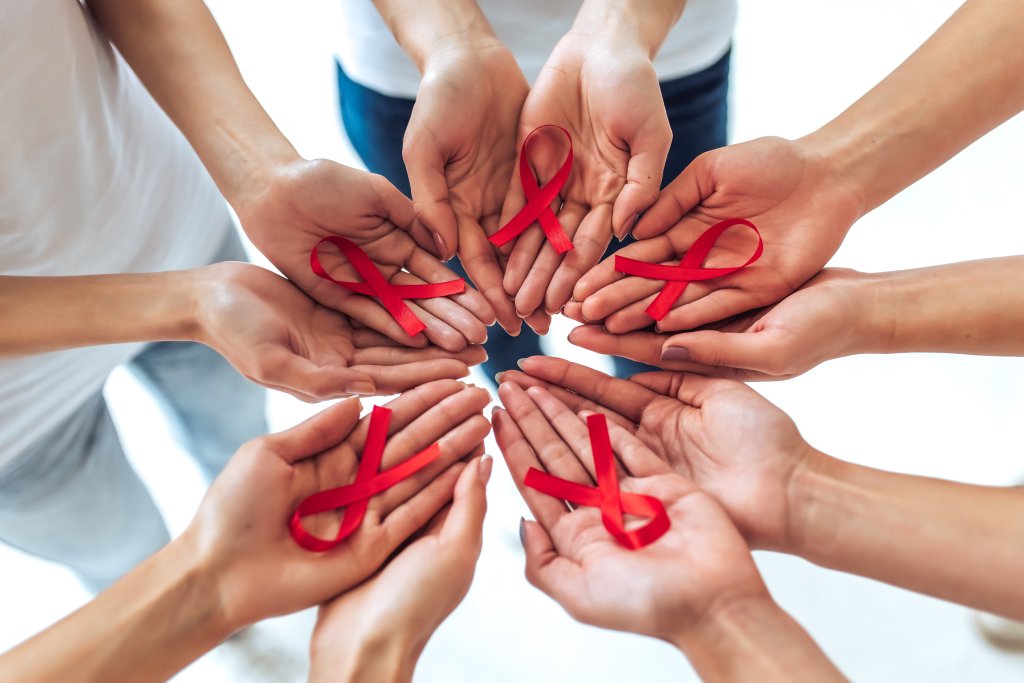 Eine Gruppe von Menschen steht im Kreis, jeder h&auml;lt ein rotes Band in der offenen Hand, als Symbol f&uuml;r die Unterst&uuml;tzung der HIV/AIDS-Aufkl&auml;rung und Solidarit&auml;t.