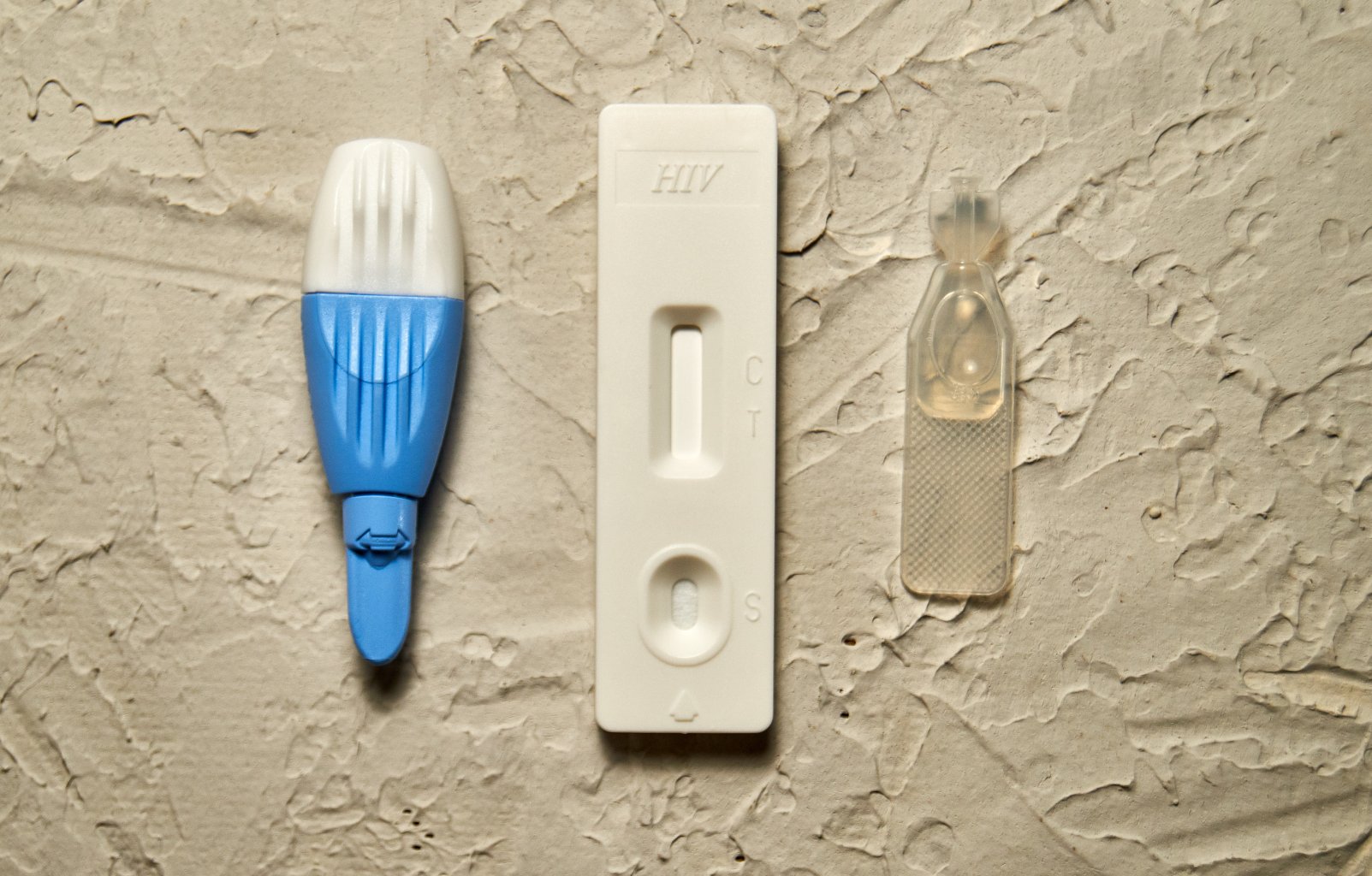 Ein HIV-Schnelltest-Kit mit drei Komponenten: einer blau-wei&szlig;en Lanzette, einer wei&szlig;en Testkassette mit der Aufschrift HIV und den mit C und T gekennzeichneten Abschnitten sowie einem kleinen durchsichtigen Probentropfer, der auf einer strukturierten Oberfl&auml;che angebracht ist.
