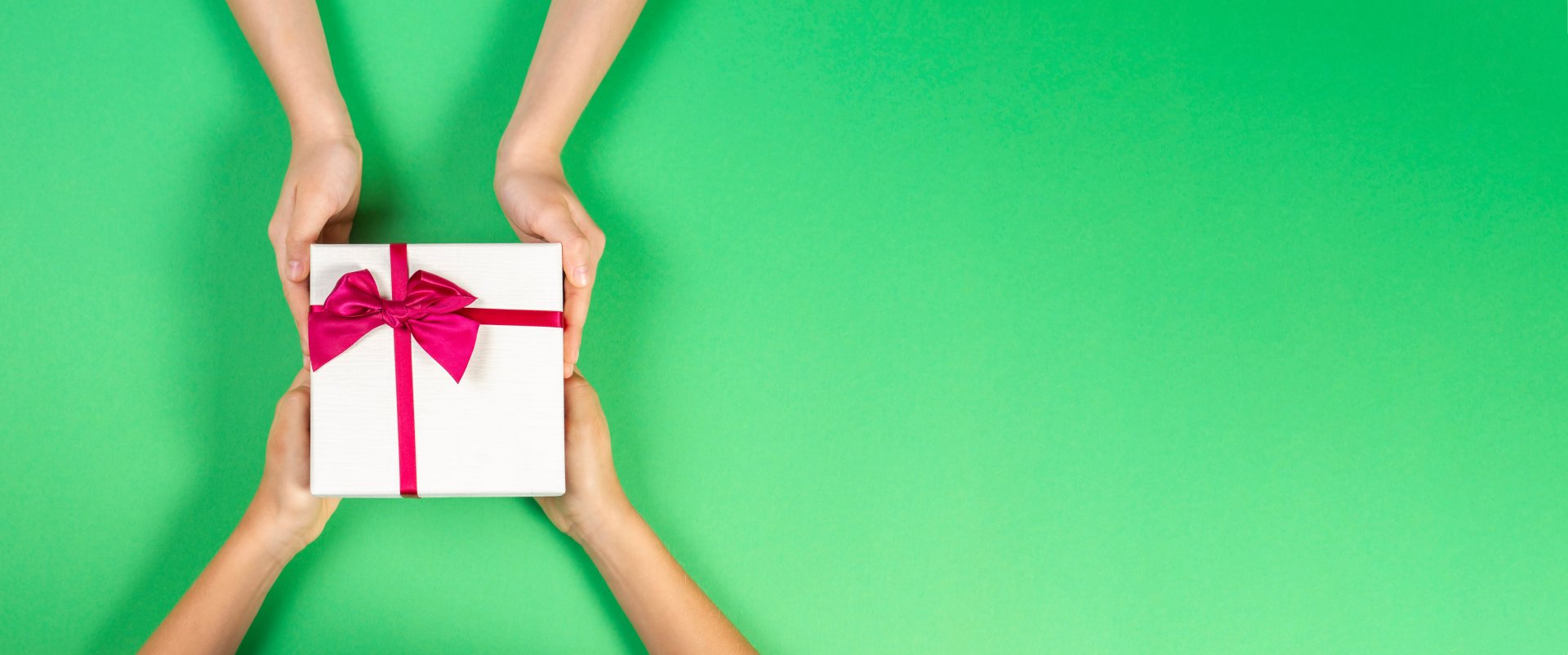 Zwei H&auml;nde &uuml;berreichen einem anderen Paar vor einem leuchtend gr&uuml;nen Hintergrund eine wei&szlig;e Geschenkschachtel mit einem rosa Band und einer Schleife, als ob sie seltene Sammelbilder verschenken w&uuml;rden.