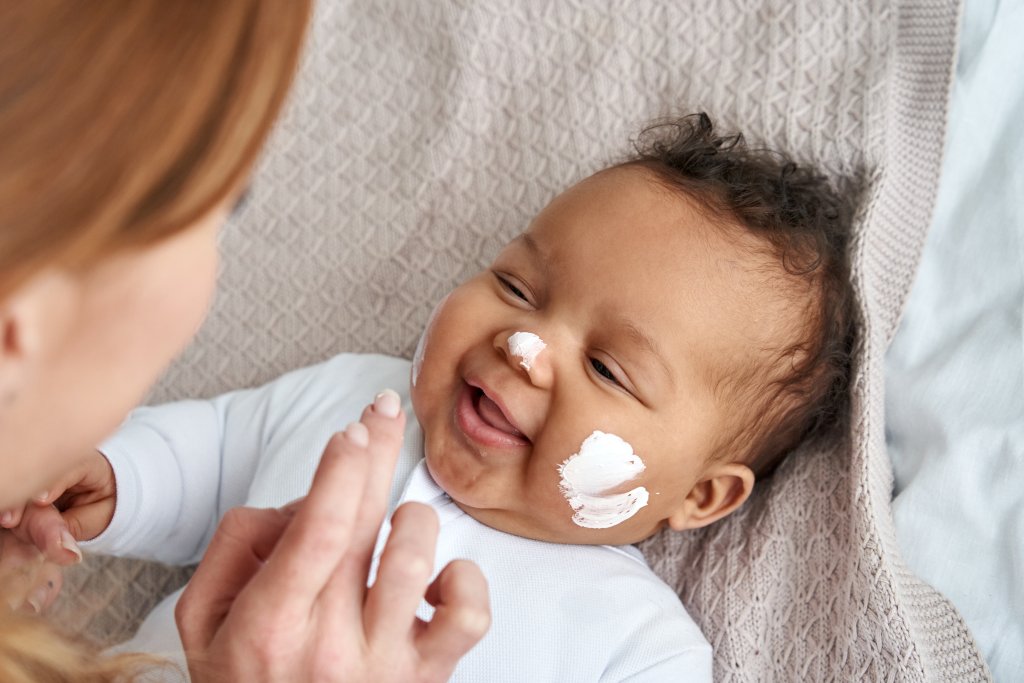 Ein l&auml;chelndes Baby auf einer Decke liegend, mit Lotion auf Wange und Nase, w&auml;hrend ein Erwachsener sanft Sonnenschutz auf das Gesicht auftr&auml;gt - so sieht Sonnenschutz bei Kindern liebevoll aus.