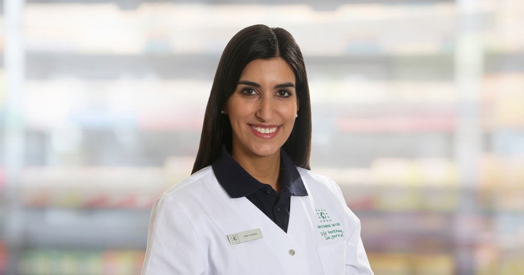 Caterina Vona, eine Frau mit langen dunklen Haaren, die einen wei&szlig;en Laborkittel und ein marineblaues Hemd mit Kragen tr&auml;gt, l&auml;chelt in die Kamera. Sie steht vor unscharfen Regalen, m&ouml;glicherweise in einer Apotheke oder einem medizinischen Bereich.