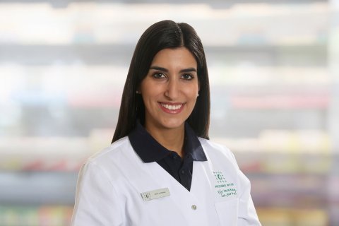 Caterina Vona, eine Frau mit langen dunklen Haaren, die einen wei&szlig;en Laborkittel und ein marineblaues Hemd mit Kragen tr&auml;gt, l&auml;chelt in die Kamera. Sie steht vor unscharfen Regalen, m&ouml;glicherweise in einer Apotheke oder einem medizinischen Bereich.