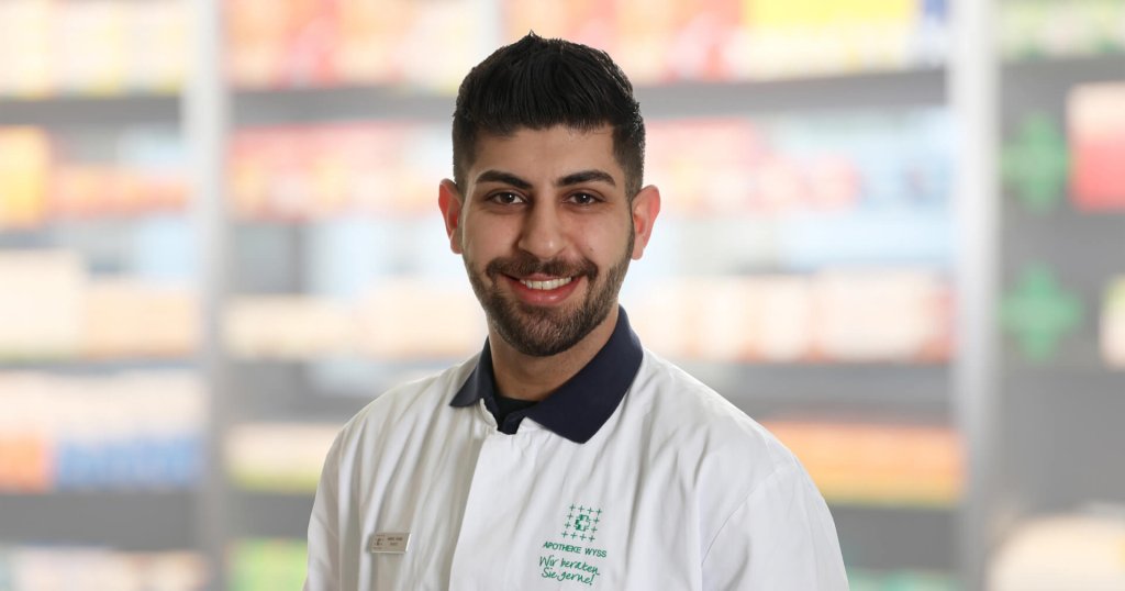 Gabriel Kouria, ein l&auml;chelnder Mann mit dunklem Haar und Bart, tr&auml;gt einen wei&szlig;en Laborkittel mit einem Namensschild und steht vor verschwommenen Apothekenregalen.