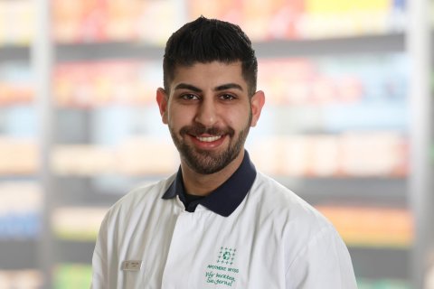 Gabriel Kouria, ein l&auml;chelnder Mann mit dunklem Haar und Bart, tr&auml;gt einen wei&szlig;en Laborkittel mit einem Namensschild und steht vor verschwommenen Apothekenregalen.