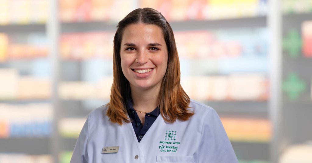 Rahel Wernli, die einen hellblauen Laborkittel mit einem Namensschild tr&auml;gt, l&auml;chelt in die Kamera. Der Hintergrund ist unscharf und zeigt Regale mit verschiedenen Produkten in einer Laden- oder Apothekenumgebung.