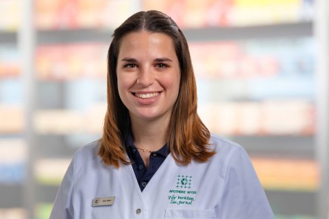 Rahel Wernli, die einen hellblauen Laborkittel mit einem Namensschild tr&auml;gt, l&auml;chelt in die Kamera. Der Hintergrund ist unscharf und zeigt Regale mit verschiedenen Produkten in einer Laden- oder Apothekenumgebung.