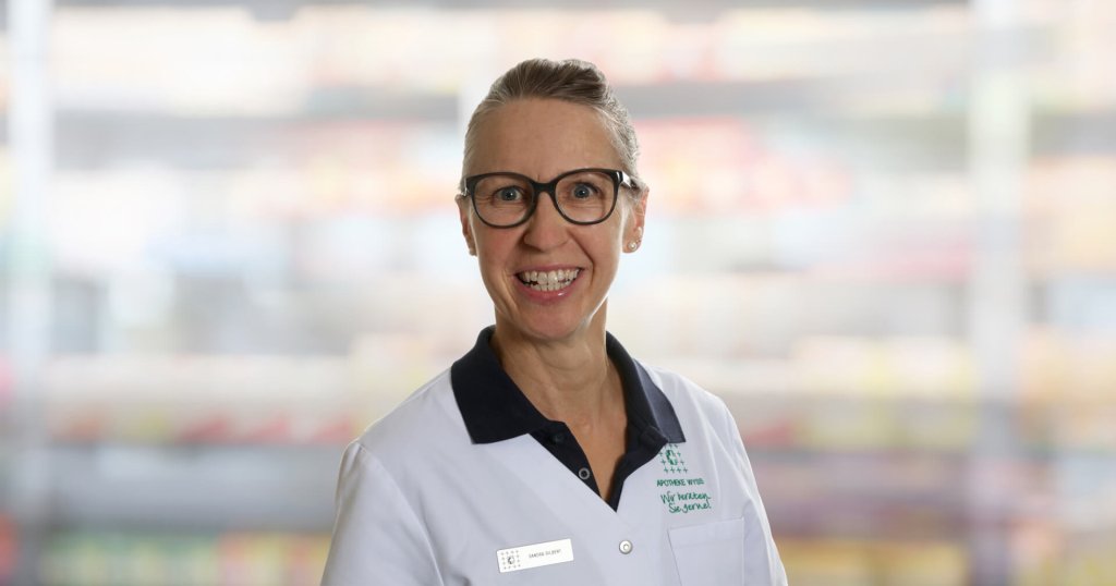 Sandra Gilbert, eine l&auml;chelnde Frau mit Brille und zur&uuml;ckgebundenem Haar, tr&auml;gt einen wei&szlig;en Laborkittel mit Namensschild und Firmenlogo und steht vor einem unscharfen Innenhintergrund.