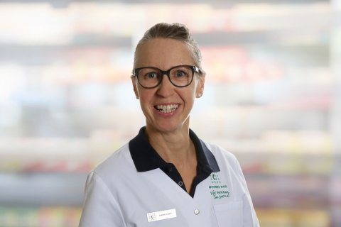 Sandra Gilbert, eine l&auml;chelnde Frau mit Brille und zur&uuml;ckgebundenem Haar, tr&auml;gt einen wei&szlig;en Laborkittel mit Namensschild und Firmenlogo und steht vor einem unscharfen Innenhintergrund.