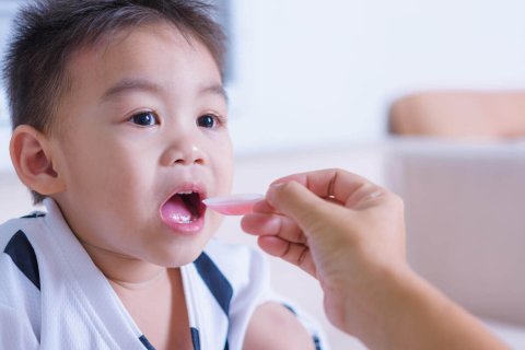 Ein kleines Kind mit kurzen Haaren &ouml;ffnet den Mund, w&auml;hrend eine erwachsene Hand ihm Medizin aus einem rosa L&ouml;ffel anbietet und die Kinderdosierungen sorgf&auml;ltig abmisst. Der Schauplatz scheint ein Haus mit einem unscharfen Hintergrund zu sein.
