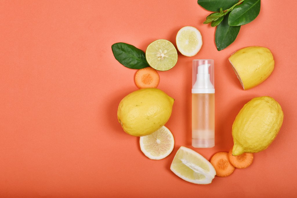 Eine Flasche Serum, umgeben von Zitronen, Limettenscheiben, Karottenscheiben und gr&uuml;nen Bl&auml;ttern auf einem orangefarbenen Hintergrund, der nat&uuml;rliche Inhaltsstoffe f&uuml;r die richtige Narbenpflege und gesunde Haut hervorhebt.