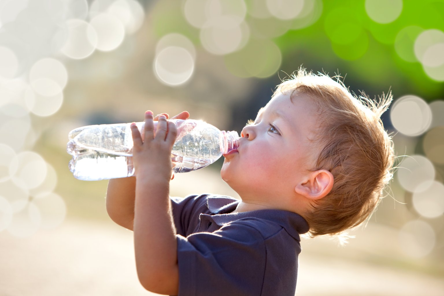 Ein kleines Kind mit hellbraunem Haar trinkt im Freien Wasser aus einer durchsichtigen Plastikflasche, um sich von der Hitze zu erholen, w&auml;hrend Sonnenlicht und ein gr&uuml;ner, verschwommener Hintergrund einen Bokeh-Effekt erzeugen.