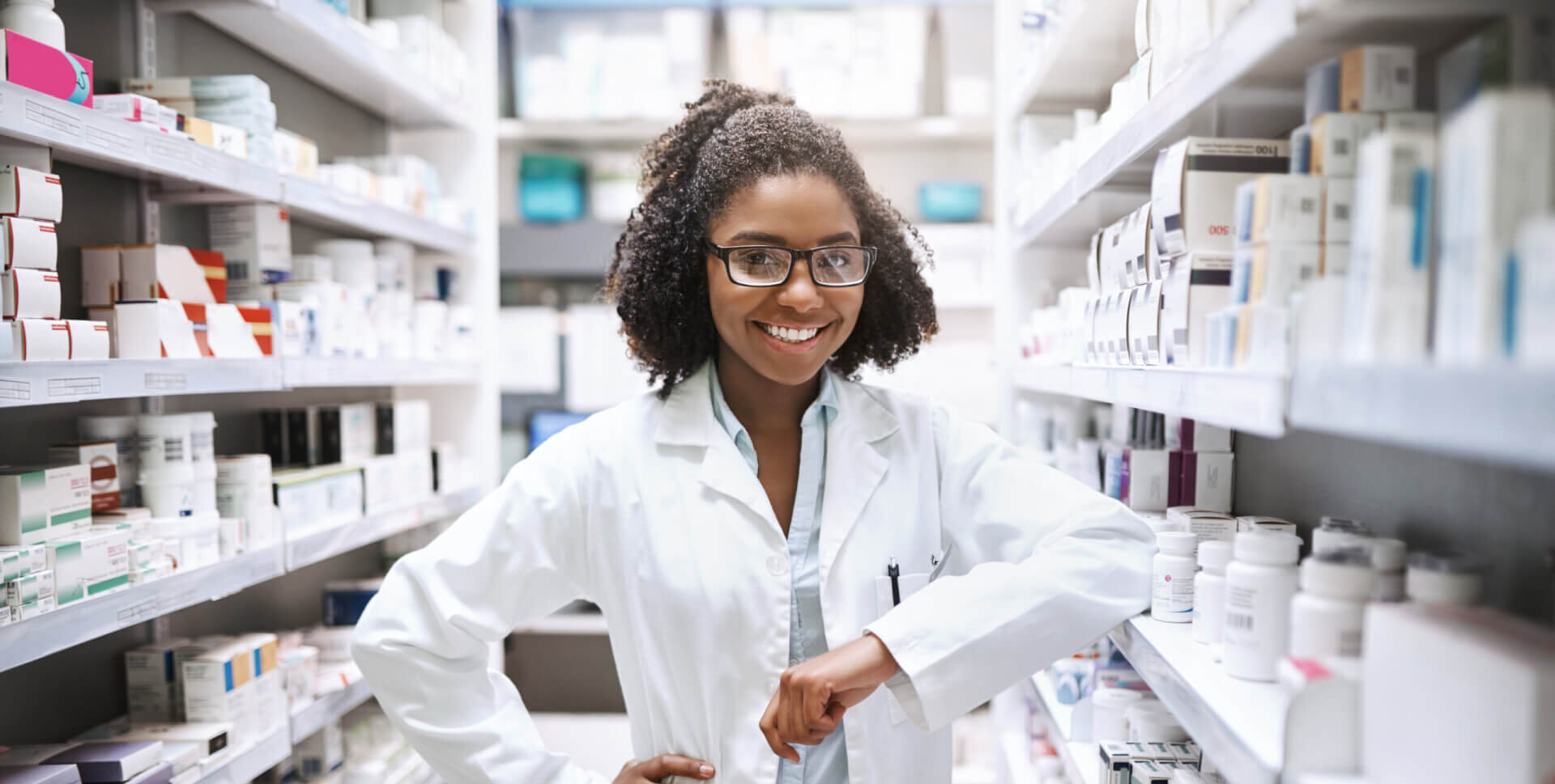 Eine l&auml;chelnde Fachfrau Apotheke EFZ im weissen Laborkittel steht selbstbewusst zwischen den mit Medikamentenflaschen und -schachteln best&uuml;ckten Apothekenregalen, den Arm auf ein Regal gest&uuml;tzt.