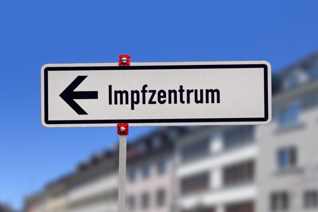 Ein Stra&szlig;enschild mit einem Pfeil nach links und dem Wort "Impfzentrum" weist den Weg zu einem COVID-19-Standort, vor einem verschwommenen Hintergrund aus Geb&auml;uden und einem klaren blauen Himmel.