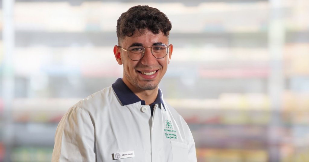 Ein l&auml;chelnder Mann mit kurzen lockigen Haaren und Brille, den sein Namensschild als Adriano Santos ausweist, tr&auml;gt einen wei&szlig;en Laborkittel und steht in einem Innenraum mit unscharfen Regalen im Hintergrund.