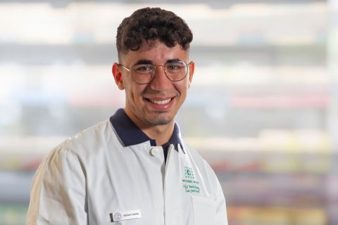 Ein l&auml;chelnder Mann mit kurzen lockigen Haaren und Brille, den sein Namensschild als Adriano Santos ausweist, tr&auml;gt einen wei&szlig;en Laborkittel und steht in einem Innenraum mit unscharfen Regalen im Hintergrund.