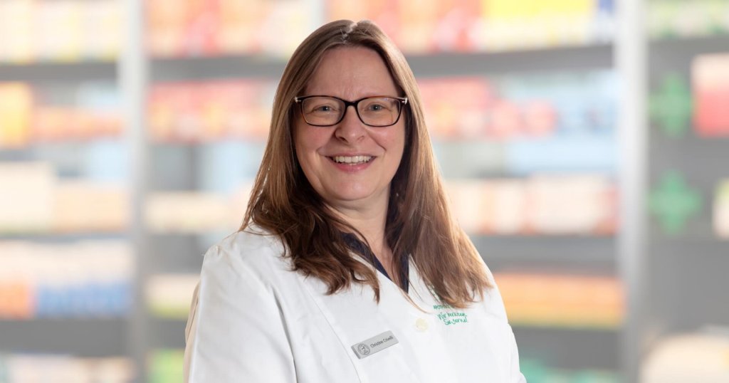 Cristina Crivelli, die eine Brille und einen wei&szlig;en Laborkittel tr&auml;gt, l&auml;chelt in die Kamera, w&auml;hrend sie vor verschwommenen Regalen mit verschiedenen Artikeln steht.