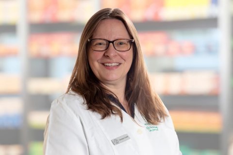 Cristina Crivelli, die eine Brille und einen wei&szlig;en Laborkittel tr&auml;gt, l&auml;chelt in die Kamera, w&auml;hrend sie vor verschwommenen Regalen mit verschiedenen Artikeln steht.