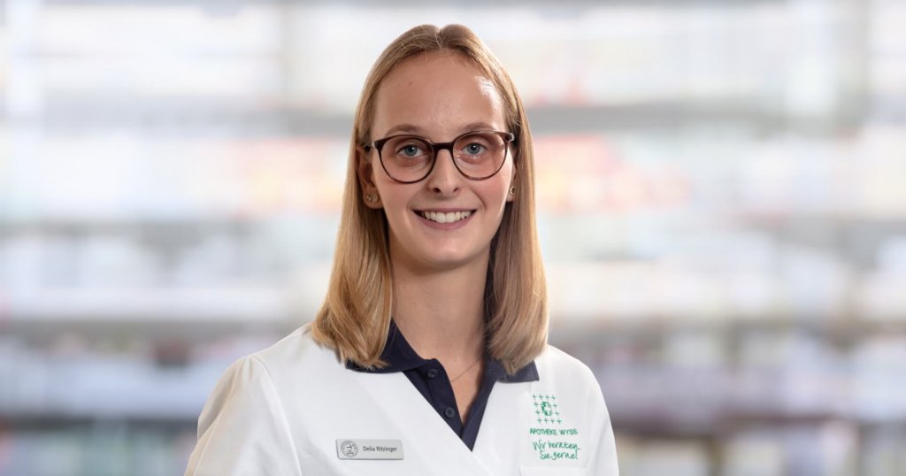 Delia Ritzinger, eine junge Frau mit glattem blondem Haar und Brille, l&auml;chelt in die Kamera. Sie tr&auml;gt einen wei&szlig;en Laborkittel und ein Namensschild und steht vor einem unscharfen Hintergrund.