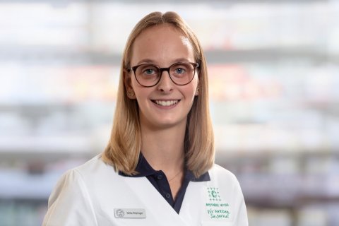 Delia Ritzinger, eine junge Frau mit glattem blondem Haar und Brille, l&auml;chelt in die Kamera. Sie tr&auml;gt einen wei&szlig;en Laborkittel und ein Namensschild und steht vor einem unscharfen Hintergrund.