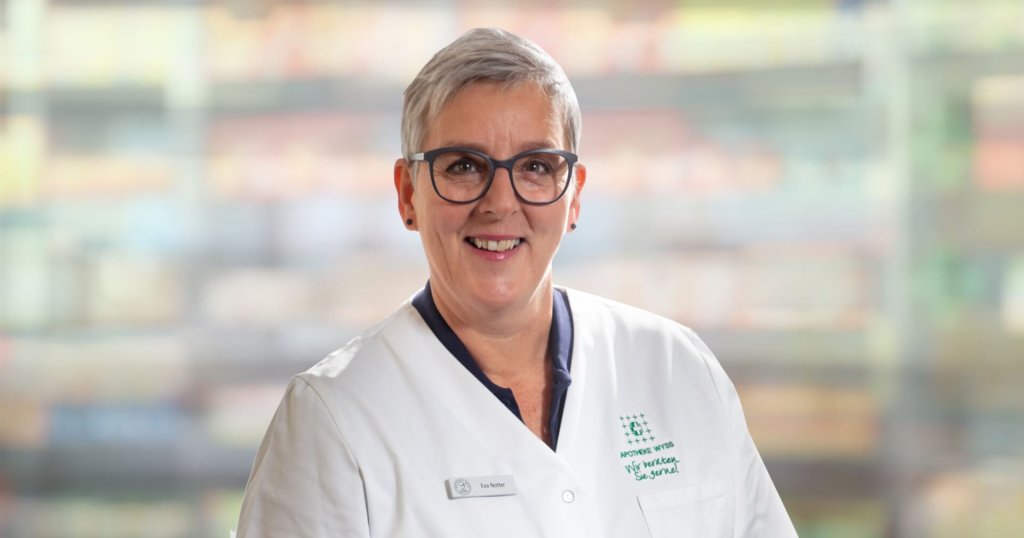 Eine l&auml;chelnde Frau mit kurzen grauen Haaren und Brille, die einen wei&szlig;en Arztkittel tr&auml;gt, steht vor einem unscharfen Hintergrund.