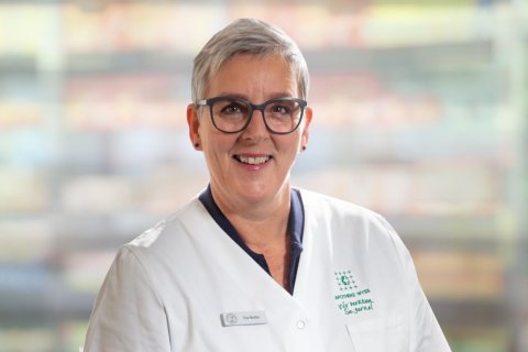 Eine l&auml;chelnde Frau mit kurzen grauen Haaren und Brille, die einen wei&szlig;en Arztkittel tr&auml;gt, steht vor einem unscharfen Hintergrund.