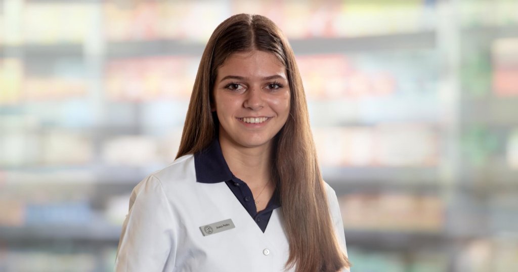 Joana Amaral Pedro, eine junge Frau mit glattem braunem Haar, tr&auml;gt einen wei&szlig;en Laborkittel und ein Namensschild, w&auml;hrend sie in die Kamera l&auml;chelt. Der unscharfe Hintergrund erinnert an ein Gesch&auml;ft oder eine Apotheke mit Regalen voller Produkte.