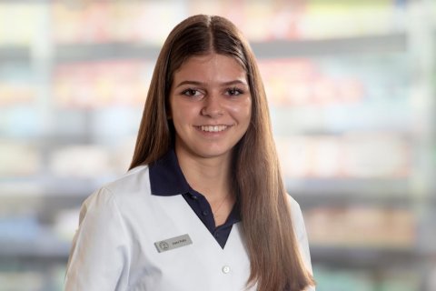 Joana Amaral Pedro, eine junge Frau mit glattem braunem Haar, tr&auml;gt einen wei&szlig;en Laborkittel und ein Namensschild, w&auml;hrend sie in die Kamera l&auml;chelt. Der unscharfe Hintergrund erinnert an ein Gesch&auml;ft oder eine Apotheke mit Regalen voller Produkte.