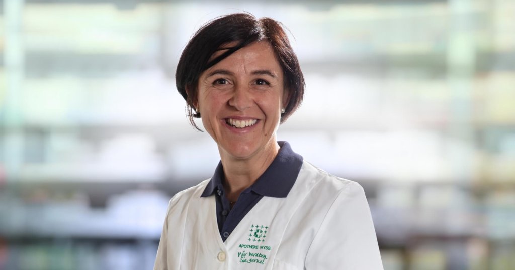 Karin Bearth Blumenthal, eine l&auml;chelnde Frau mit kurzen dunklen Haaren, tr&auml;gt einen wei&szlig;en Laborkittel &uuml;ber einem marineblauen Kragenhemd und steht vor einem unscharfen Hintergrund, der an Regale in einer Apotheke oder einem Labor erinnert.