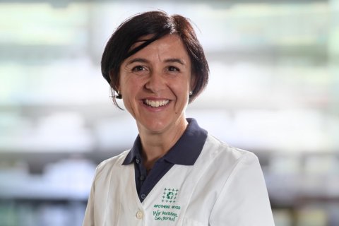 Karin Bearth Blumenthal, eine l&auml;chelnde Frau mit kurzen dunklen Haaren, tr&auml;gt einen wei&szlig;en Laborkittel &uuml;ber einem marineblauen Kragenhemd und steht vor einem unscharfen Hintergrund, der an Regale in einer Apotheke oder einem Labor erinnert.