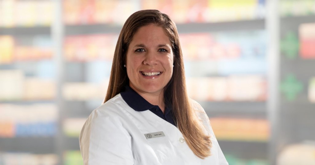 Eine Frau mit langen braunen Haaren, die einen wei&szlig;en Laborkittel und ein Namensschild tr&auml;gt, l&auml;chelt in die Kamera. Der Hintergrund scheint aus Regalen mit Produkten zu bestehen, wahrscheinlich in einer Apotheke oder einem Gesch&auml;ft.