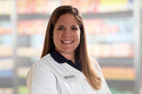 Eine Frau mit langen braunen Haaren, die einen wei&szlig;en Laborkittel und ein Namensschild tr&auml;gt, l&auml;chelt in die Kamera. Der Hintergrund scheint aus Regalen mit Produkten zu bestehen, wahrscheinlich in einer Apotheke oder einem Gesch&auml;ft.