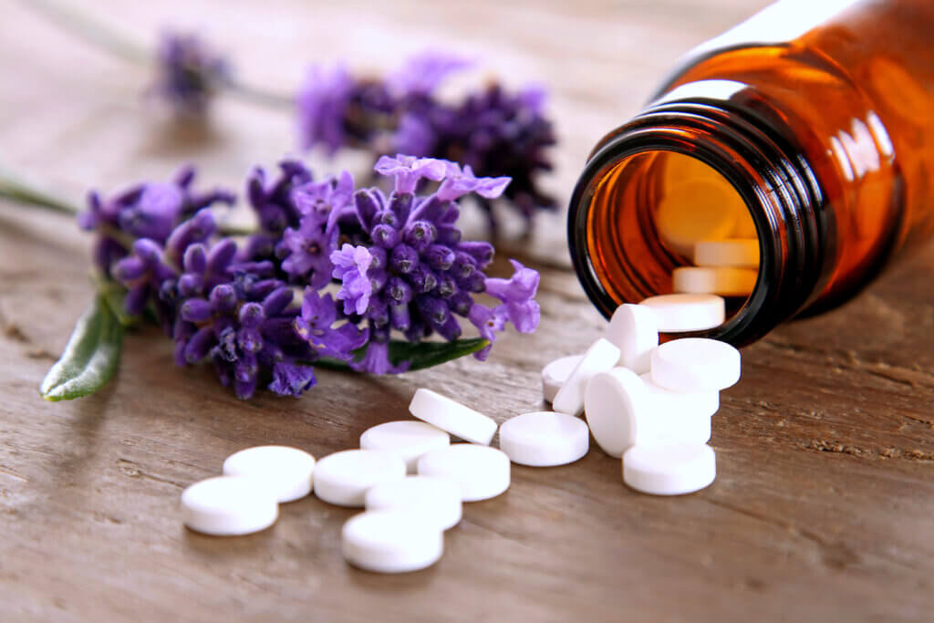 Eine bernsteinfarbene Pillenflasche mit Sch&uuml;ssler Salz ist umgekippt und die wei&szlig;en Tabletten liegen auf einer h&ouml;lzernen Fl&auml;che neben frischen Zweigen mit lila Lavendelbl&uuml;ten.