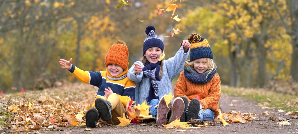 Drei kleine Kinder mit bunten Strickm&uuml;tzen und Pullovern sitzen auf einem mit Laub bedeckten Weg und lachen, w&auml;hrend sie Herbstbl&auml;tter in die Luft werfen, umgeben von B&auml;umen mit Herbstlaub - ein fr&ouml;hlicher Moment, der das Immunsystem st&auml;rken und die Gesundheit f&ouml;rdern kann.