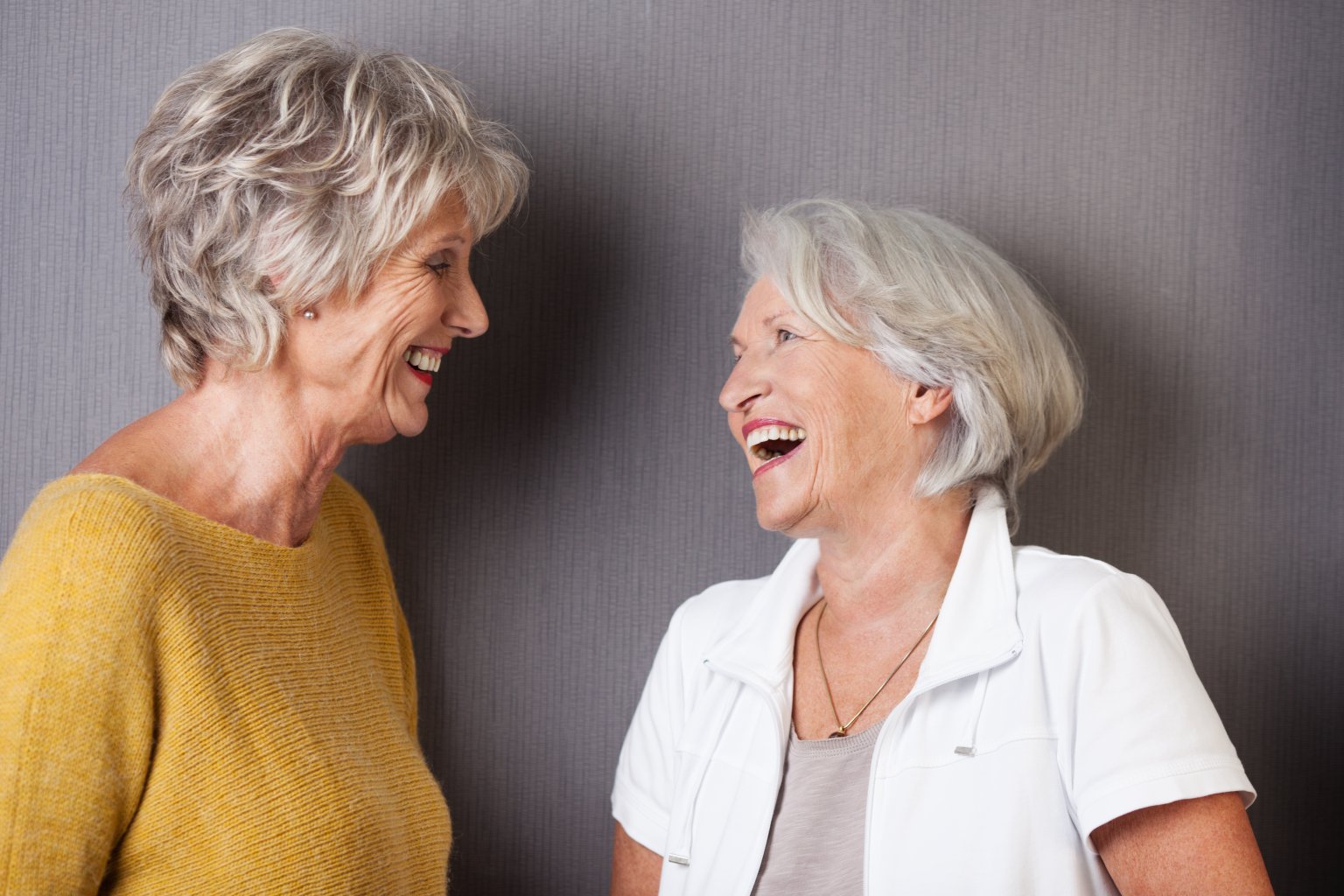 Zwei &auml;ltere Frauen mit kurzen grauen Haaren stehen und lachen zusammen - Lachen, das nachweislich das Immunsystem st&auml;rkt. Die eine tr&auml;gt einen gelben Pullover, die andere ein wei&szlig;es Oberteil. Sie stehen sich vor einem grau strukturierten Hintergrund gegen&uuml;ber.
