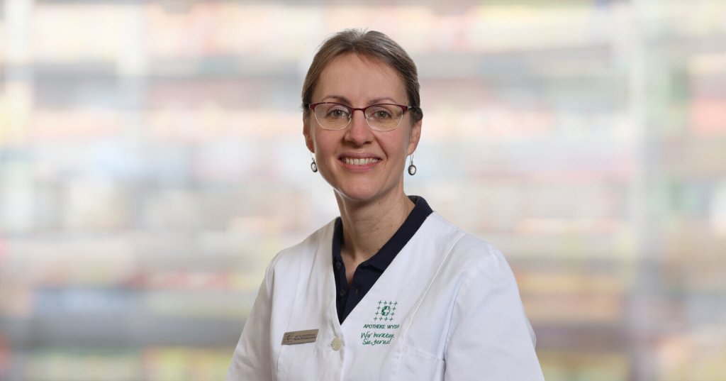 Eine Frau mit Brille und wei&szlig;em Laborkittel, Fach&auml;rztin f&uuml;r Alternativmedizin, l&auml;chelt in die Kamera. Sie hat ihr hellbraunes Haar zur&uuml;ckgebunden und steht vor einem unscharfen Hintergrund.