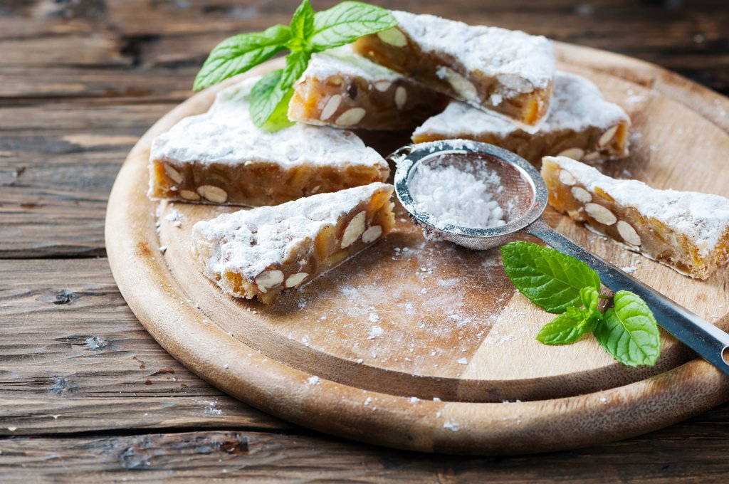 Aufgeschnittene Nuss- und Trockenfr&uuml;chtekuchen mit Puderzucker best&auml;ubt auf einem Holzbrett, begleitet von einem Metallsieb und frischen Minzbl&auml;ttern, pr&auml;sentieren die hausgemachten Leckereien auf einer rustikalen Holzoberfl&auml;che.