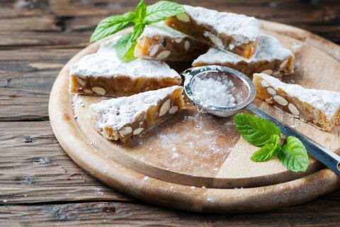 Aufgeschnittene Nuss- und Trockenfr&uuml;chtekuchen mit Puderzucker best&auml;ubt auf einem Holzbrett, begleitet von einem Metallsieb und frischen Minzbl&auml;ttern, pr&auml;sentieren die hausgemachten Leckereien auf einer rustikalen Holzoberfl&auml;che.