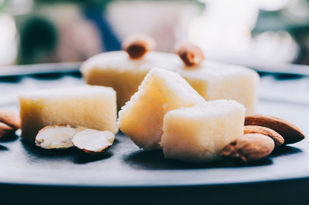 Nahaufnahme von indischen S&uuml;&szlig;igkeitenw&uuml;rfeln, wahrscheinlich aus Mandeln oder Cashewkernen, auf einem dunklen Teller mit ganzen und halbierten Mandeln - perfekte Leckereien f&uuml;r jede Gelegenheit. Der Hintergrund ist unscharf.