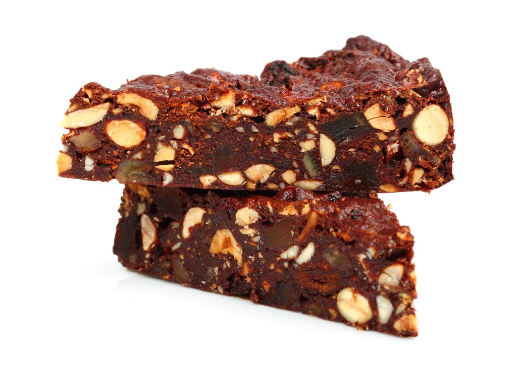 Zwei Scheiben dunkler, dichter Frucht- und Nusskuchen auf wei&szlig;em Grund, bei denen Mandeln und getrocknete Fr&uuml;chte in den reichhaltigen, feuchten Leckereien zu sehen sind - sie erinnern an Leckereien aus einer alten Apotheke.