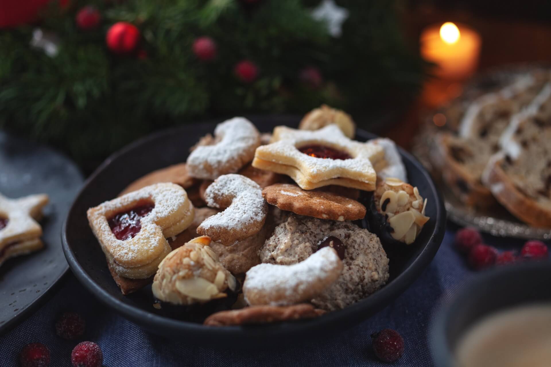 Eine schwarze Schale mit verschiedenen Leckereien, darunter herz- und sternf&ouml;rmige Kekse, die mit Puderzucker best&auml;ubt sind, steht auf einem Tisch mit festlicher Dekoration und einer brennenden Kerze im Hintergrund.