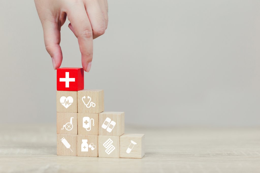 Eine Hand stapelt Holzbl&ouml;cke mit medizinischen Symbolen und legt einen roten Block mit einem wei&szlig;en Kreuz darauf, der die Notfallapotheke symbolisiert, die Tag und Nacht zur Verf&uuml;gung steht. Die Bl&ouml;cke stehen f&uuml;r den Aufbau der medizinischen Grundversorgung und die medizinische Notfallversorgung.