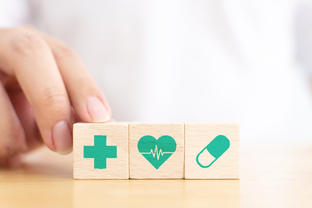 Eine Hand ordnet drei Holzbl&ouml;cke auf einem Tisch an, auf denen jeweils ein gr&uuml;nes Symbol zu sehen ist - ein medizinisches Kreuz, ein Herz mit einer Herzschlaglinie und eine Kapselpille -, die Gesundheitskonzepte wie die Tag und Nacht verf&uuml;gbaren Dienste der Notfallapotheke symbolisieren.