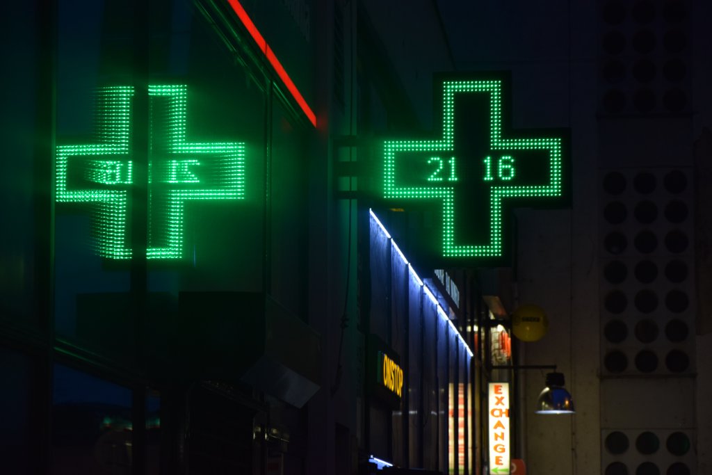 Ein gr&uuml;n leuchtendes digitales Notfallapotheken-Kreuz zeigt nachts die Uhrzeit "21:16" an, deren Spiegelung auf einer nahen Glasfl&auml;che sichtbar ist. Tag und Nacht erhellen rote und blaue Lichter den Hintergrund und betonen die st&auml;ndige Verf&uuml;gbarkeit.