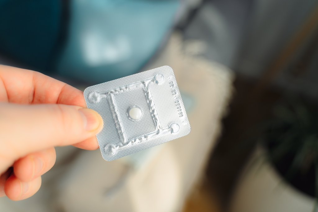 Eine Hand h&auml;lt eine Blisterpackung mit einer einzelnen wei&szlig;en Pille, wahrscheinlich aus einer Notfallapotheke, vor einem unscharfen Innenraumhintergrund - m&ouml;glicherweise ein Notfallkontrazeptivum f&uuml;r den Tag und die Nacht.