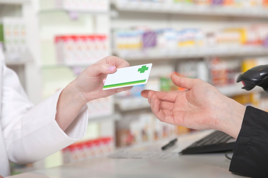 Ein Apotheker reicht einem Kunden eine Apothekenkarte &uuml;ber einen Tresen in einer Notfallapotheke, im Hintergrund stehen Regale mit Medikamenten. Der Schwerpunkt liegt auf dem Austausch der Karte, der den Tag- und Nachtservice f&uuml;r den st&auml;ndigen Zugang der Kunden zur Versorgung hervorhebt.