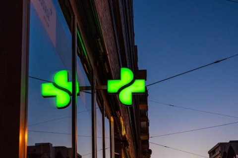 Ein leuchtend gr&uuml;nes Apothekenkreuz mit der Aufschrift "Tag und Nacht" ist in der Abendd&auml;mmerung an einem Geb&auml;ude angebracht und spiegelt sich im Fenster. Stromleitungen und Silhouetten von Geb&auml;uden heben sich vom Abendhimmel im Hintergrund ab.