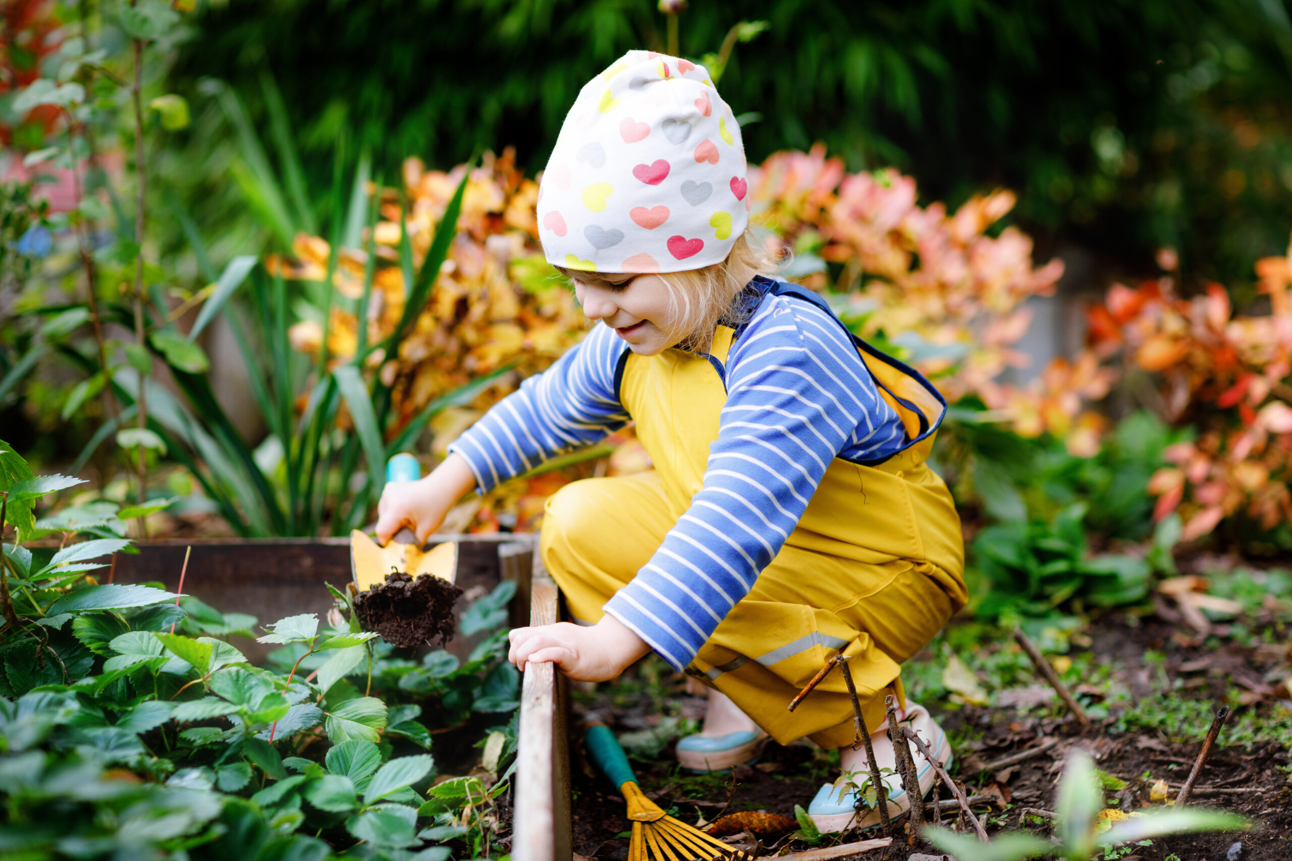 Ein kleines Kind mit herzf&ouml;rmigem Hut und gelber Latzhose gr&auml;bt mit einer kleinen Schaufel in einem Gartenbeet und zeigt, dass G&auml;rtnern eine spielerische Form der Therapie inmitten von gr&uuml;nen Pflanzen und buntem Laub sein kann.