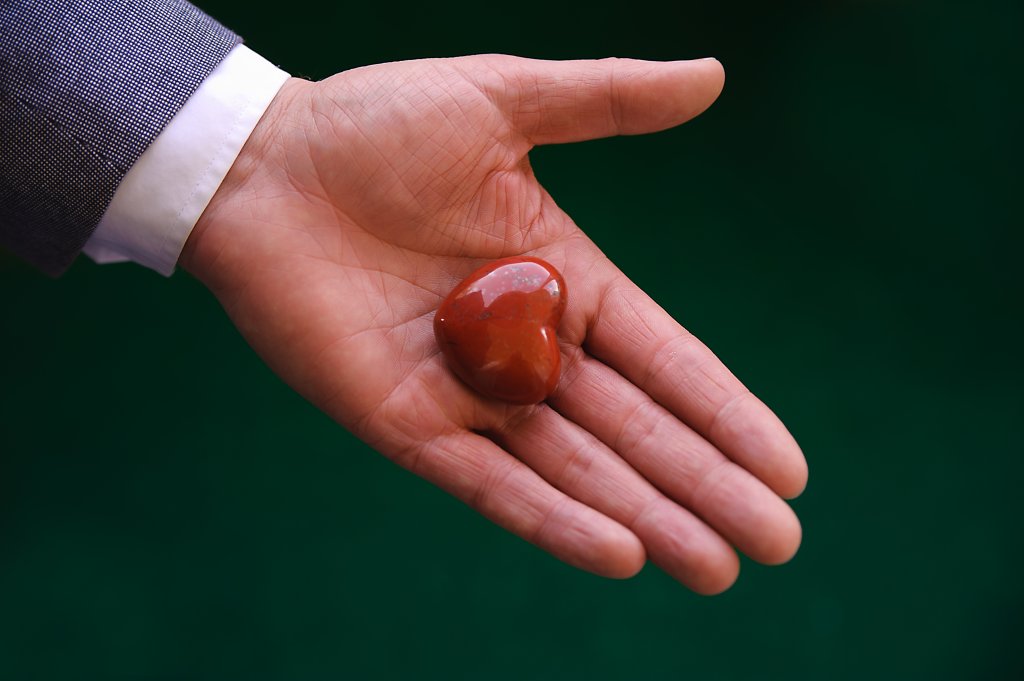 Eine Person h&auml;lt mit der offenen Hand einen geschliffenen, herzf&ouml;rmigen roten Stein (Herz) vor einem dunkelgr&uuml;nen Hintergrund. Die Person tr&auml;gt eine blaue Jacke und ein wei&szlig;es Hemd.